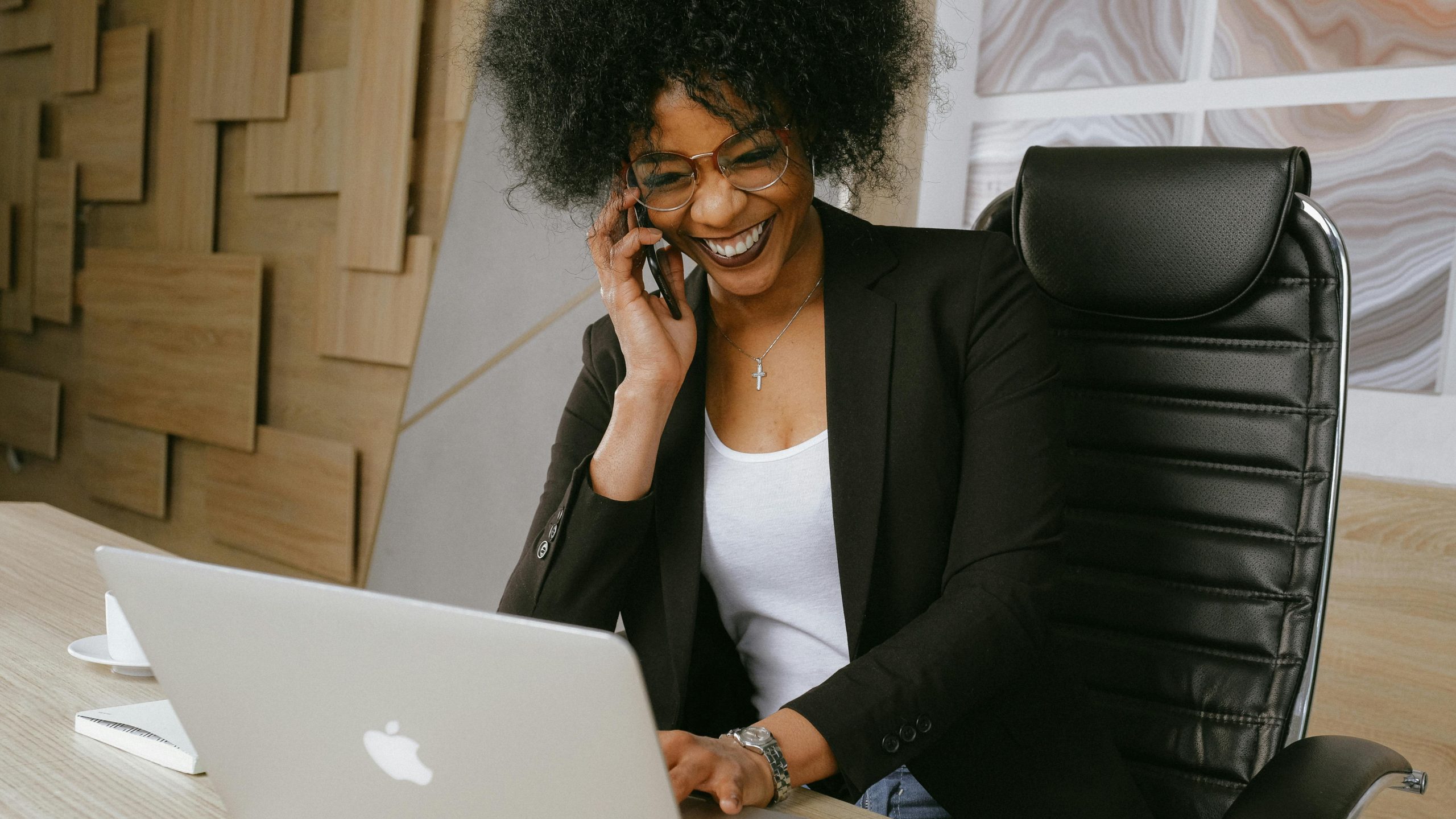 Smiling woman on phone call while working on a laptop in a stylish office.